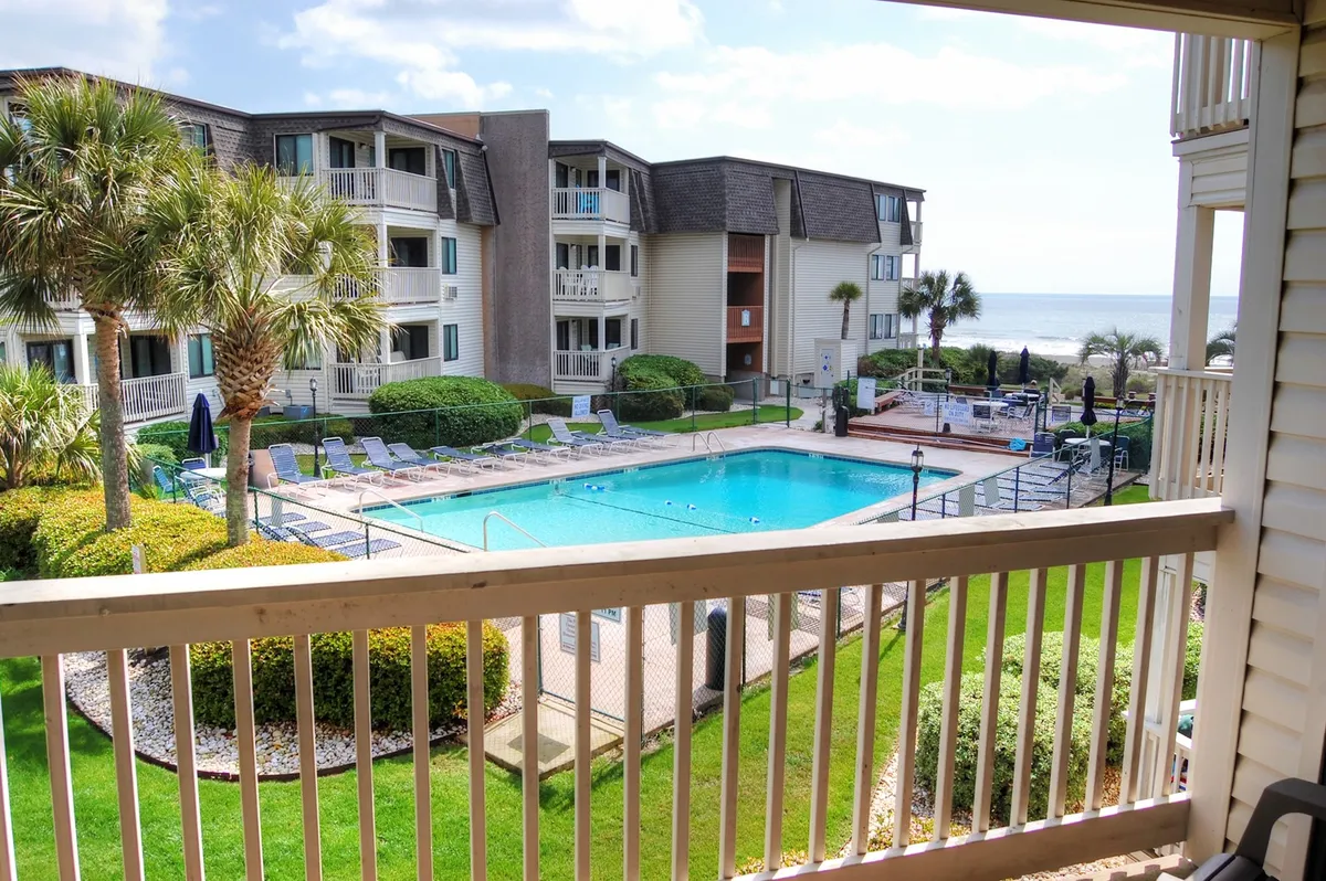 A sparkling outdoor swimming pool surrounded by palm trees and landscaped gardens, with balconies overlooking the pool. The scene is tranquil and inviting, perfect for both family activities and quiet relaxation. There are hints of barbecuing and gathering areas, adding to the communal feel.