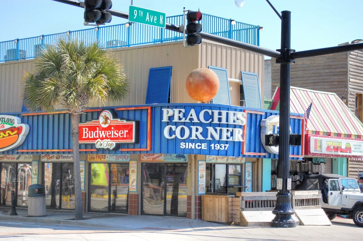 A lively Myrtle Beach streetscape featuring the iconic Peaches Corner sign, colorful storefronts, and bustling street life. The mood is festive and social, highlighting opportunities for dining, live music, and people-watching in the heart of the entertainment district.