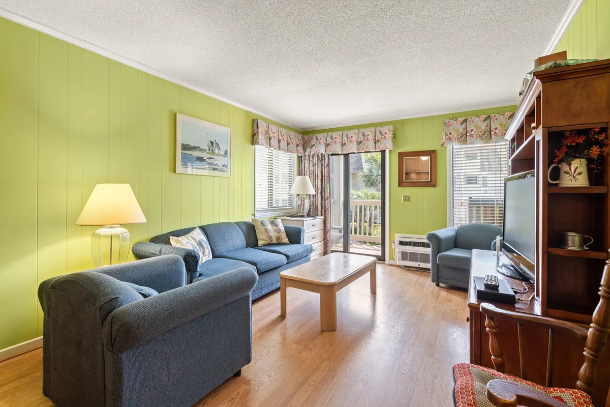 This interior shot highlights a living area with comfortable seating grouped around a wooden coffee table, green accent walls, and a large window overlooking an outside view. The natural light streaming in evokes the idea of sunny mornings and cozy afternoons, while the arrangement encourages conversation and relaxation.