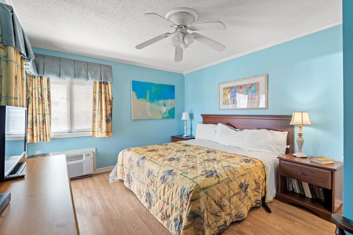A spacious, comfortable bedroom featuring a king bed with a floral quilt, a bedside lamp, framed artwork, and a TV on the dresser. Natural light softly fills the room, creating a serene and private ambiance.