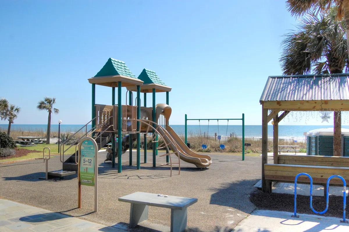 A cheerful playground scene overlooking the ocean, complete with swings, a colorful play structure, and picnic areas where families and children gather. The backdrop of the sea and sand adds a distinctly Myrtle Beach feel, emphasizing the joy of outdoor family fun.
