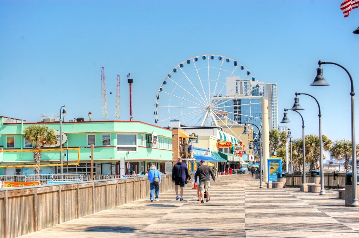 A vibrant scene along Myrtle Beach's iconic boardwalk, featuring a large Ferris wheel towering over colorful storefronts and a lively crowd of people strolling, shopping, and enjoying the various attractions. The boardwalk and amusements create an exciting, festive atmosphere, perfect for families.