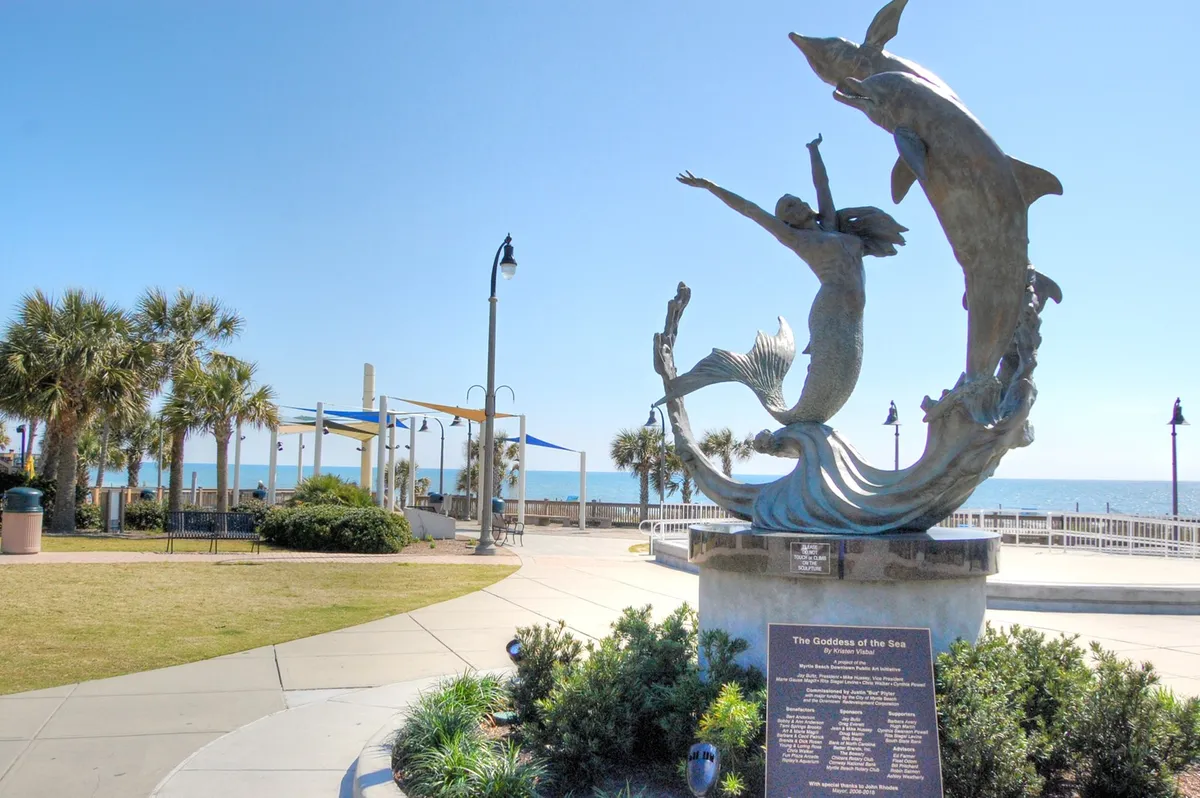A vibrant exterior scene featuring the iconic Goddess of the Sea sculpture, with the ocean and boardwalk in the background. Palm trees line the area, and people are seen walking, enjoying the fresh air, and engaging with local art.