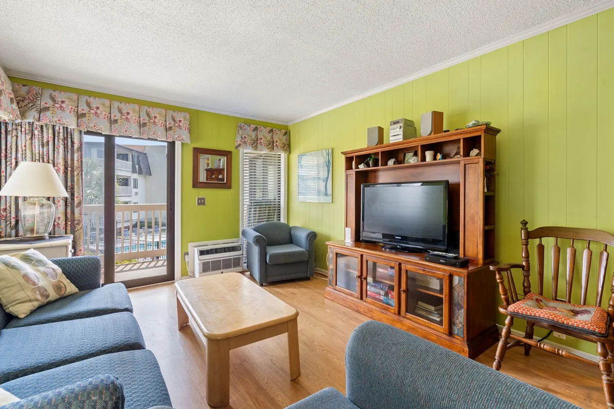 A welcoming living room setup featuring a comfortable sofa (potential sleeper sofa), a television, wooden cabinet, and light curtains framing a sunlit window. The space is ideal for relaxing or socializing after a long day.