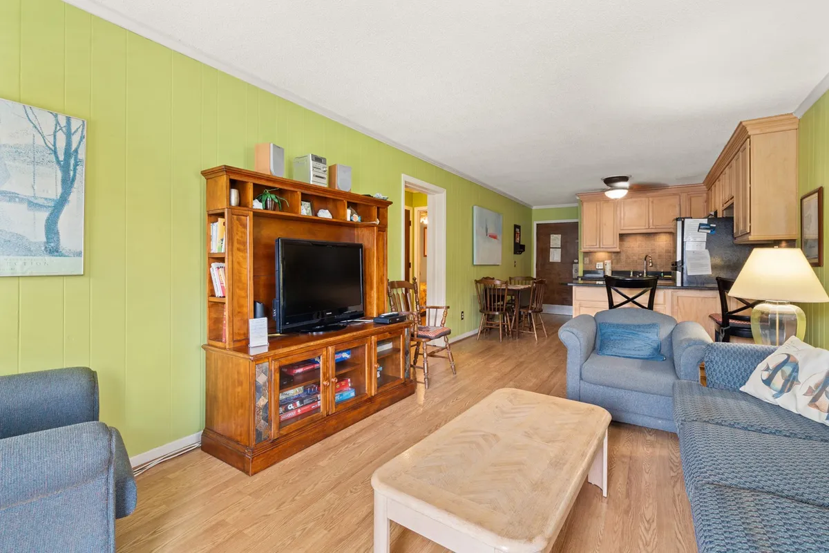 This image presents an open-plan living space featuring a comfortable sofa, TV unit, dining area, and a visible entrance to the kitchen. The space appears inviting and cohesive, encouraging gatherings and shared experiences. The layout allows for easy movement between social and dining zones, and the lighting suggests a bright and welcoming atmosphere.