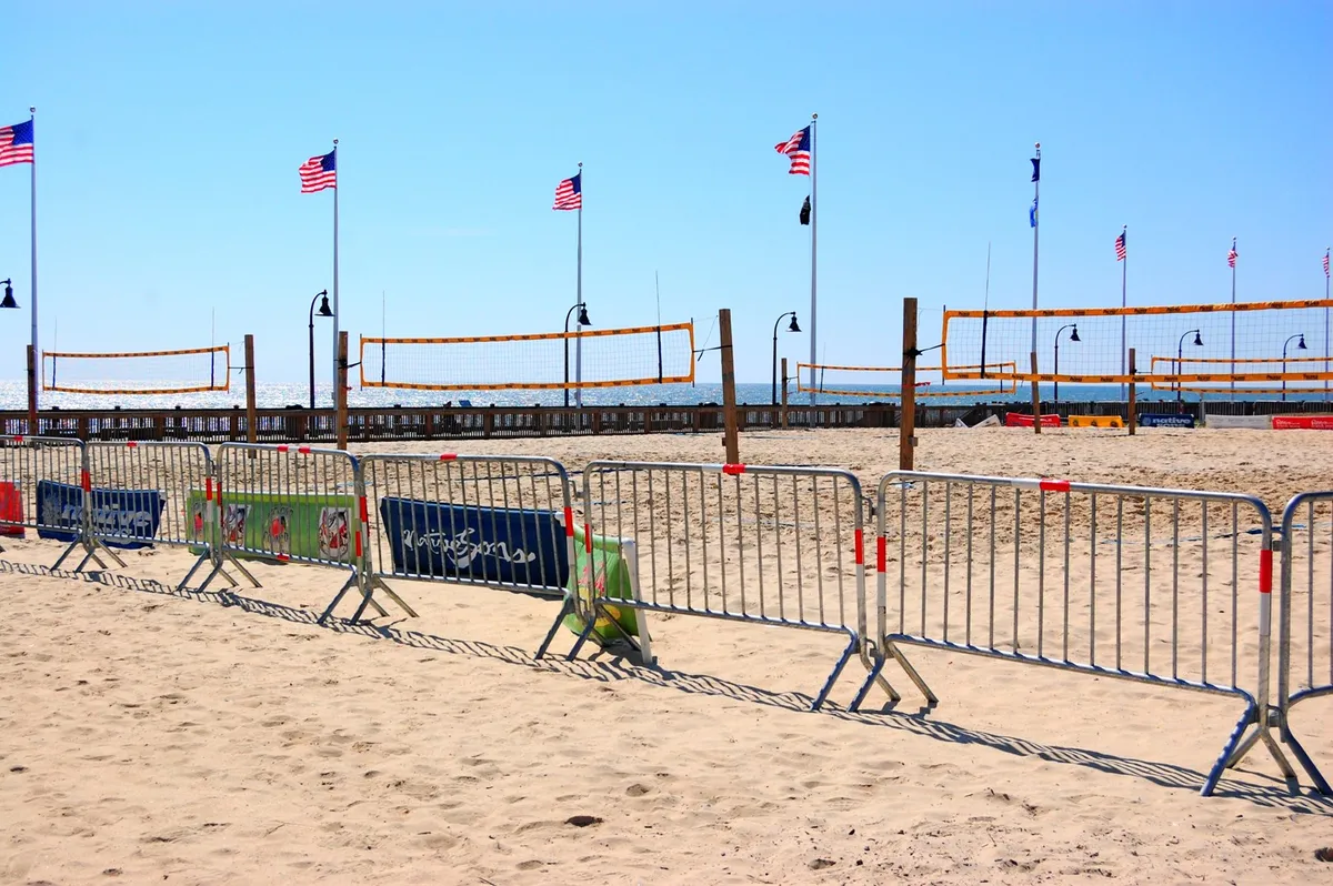 This lively exterior scene captures a bustling beachside volleyball tournament, with nets set up on the sand, American flags waving in the background, and groups of people sunbathing and strolling along the shore. The golden sunset bathes the entire area in a warm, energetic glow, suggesting endless opportunities for outdoor fun and relaxation just steps from the property.