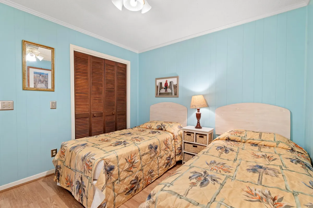 A bright second bedroom with twin beds, each covered in floral bedspreads. A wooden nightstand with a lamp sits between the beds, with framed artwork on the wall and sunlight streaming in through the window. There's a closet with wooden doors, offering ample storage.