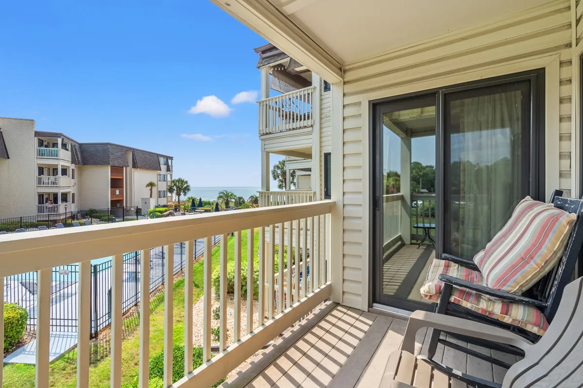 The image showcases the exterior of a beachfront condo in Myrtle Beach, highlighting a private balcony with sweeping ocean views, a sparkling pool area surrounded by palm trees, and the inviting ambiance of the Golden Mile. The scene evokes relaxation, with guests unwinding on the balcony, enjoying morning coffee, or socializing by the pool as the sun rises over the Atlantic.