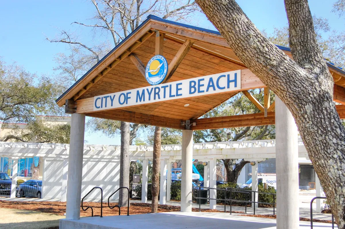An exterior shot focusing on the 'City of Myrtle Beach' signage set against a wooden structure and surrounded by lush trees. The welcoming area is ideal for informal community gatherings or learning about local events.